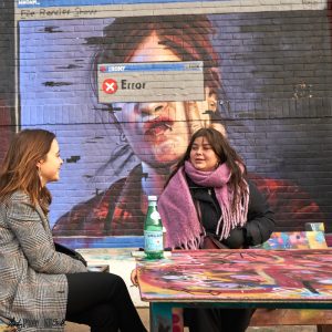 Two women drinking posh bottled water in front of an irony graffito